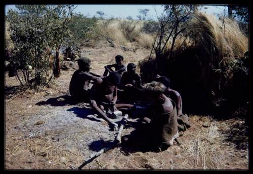 Groups: Men sitting in front of a skerm, including !Naishi of Band 5 wearing a hat, "Old Demi" of Band 6 (left) and "/Gao Music" pounding something with a metal tool on a stone