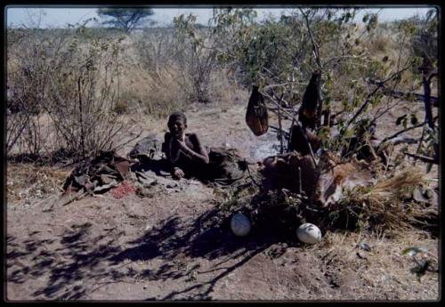 Groups: Khwo//o (mother of "/Gao Music") lying on a kaross in her dwelling place without a skerm, with a water bag made from an animal stomach hanging on a bush and ostrich eggshells and veldkos on the ground