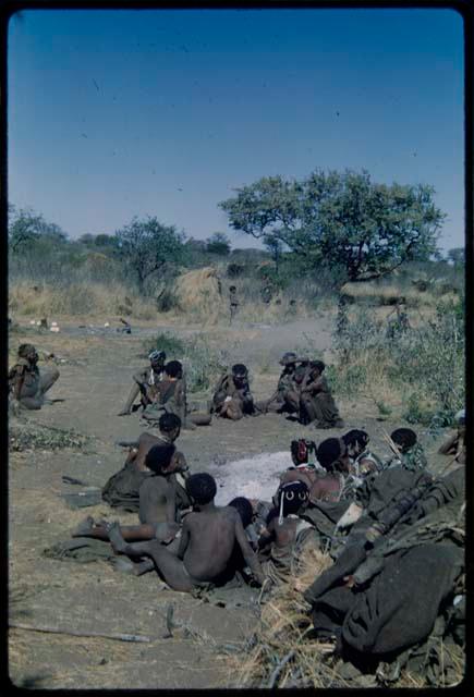 Groups: People sitting around the ashes of "Gao Medicine's" fire; showing ant hill, N!ai and /Gunda's wedding skerm, and a tree in a dance circle in the background