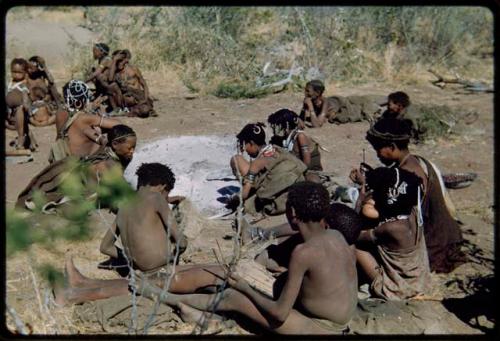 Groups: Group of people sitting around the ashes of "Gao Medicine's" fire, including a boy playing a //guashi, //Khuga (daughter of "Gao Medicine") wearing a brown headband, Khwo//o (right background), !U wearing ornaments, !Ungka (≠Toma's sister), Di!ai (left, leaning forward), and Be and Khuan//a in the background (left)