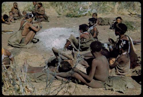 Groups: Group of people sitting around the ashes of "Gao Medicine's" fire, including a boy playing a //guashi, //Khuga (daughter of "Gao Medicine") wearing a brown headband, Khwo//o (right background), !U wearing ornaments, !Ungka (≠Toma's sister), and Be and Khuan//a in the background (left)