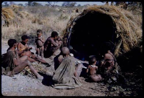 Groups: People sitting in front of Gau's skerm, including "/Qui Hunter" (inside the skerm), "Old Gau" squatting by the entrance, //Khuga (wife of !Naishi), /Naoka, "Old /Gasa" with shaved head, /Qui (son of Gau and Khuan//a), and Be (Gau's first wife)