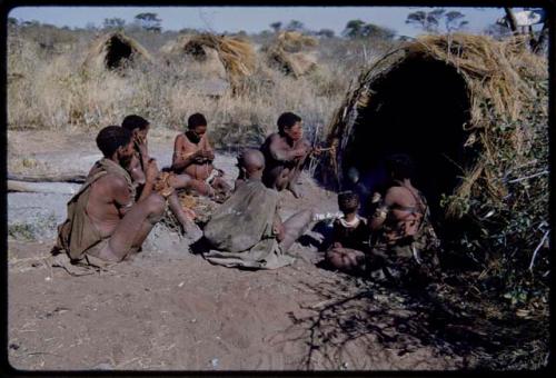 Groups: People sitting in front of Gau's skerm, including "/Qui Hunter" (inside the skerm), "Old Gau" squatting by the entrance, //Khuga (wife of !Naishi), /Naoka, "Old /Gasa" with shaved head, /Qui (son of Gau and Khuan//a), and Be (Gau's first wife)