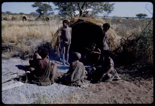 Groups: People sitting in front of Gau's skerm, including "/Qui Hunter" tasting the broth he is cooking, two boys standing, /Naoka, "Old /Gasa" with shaved head, and Be (Gau's first wife)