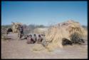 Groups, Extended Family: /Naoka and a child walking toward a group of people sitting in front of Gau's skerm, with a kudu head on the ground next to the skerm, view from behind