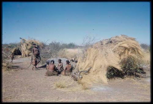 Groups, Extended Family: /Naoka and a child walking toward a group of people sitting in front of Gau's skerm, with a kudu head on the ground next to the skerm, view from behind