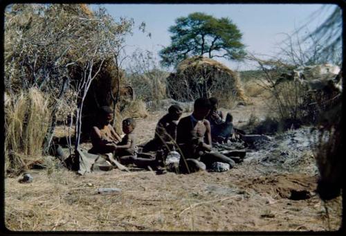 Groups, Extended Family: Gau hammering something with a stone, sitting with his family in front of a skerm