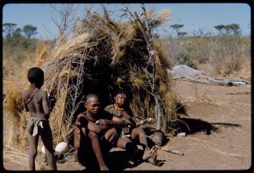 Groups, "Nuclear Family": "/Qui Navel" and //Kushay sitting in front of their skerm, with their son, N!aishi, standing next to them