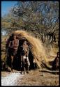 Groups, "Nuclear Family": Tsamgao and Bau (daughter of /Ti!kay) standing in front of a skerm, with an unidentified woman sitting next to them