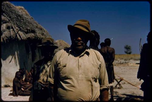 William Hartley standing, with other people sitting and standing next to a hut behind him