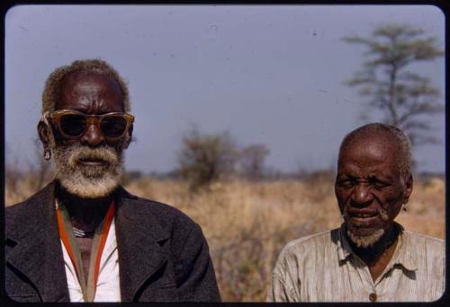 Shikongo, Ovambo headman in Mulungo, wearing dark glasses, standing with an unidentified man