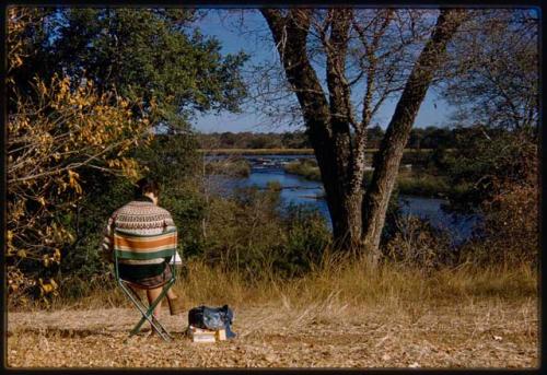 Deborah Marshall sitting in a chair and sketching by river, seen from behind