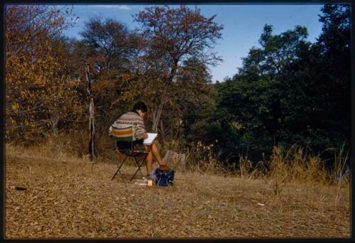 Deborah Marshall sitting in a chair and sketching by river, seen from behind