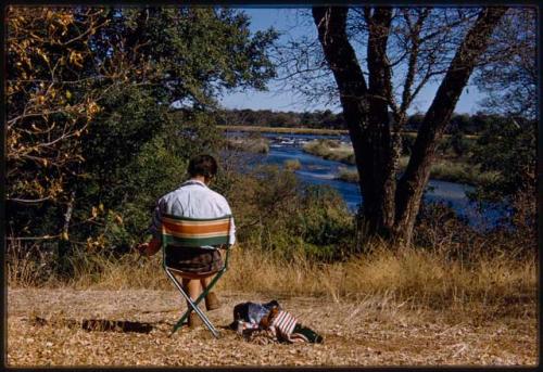 Deborah Marshall sitting in a chair and sketching by river, seen from behind