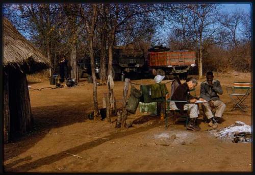 Lorna Marshall and Kernel Ledimo sitting in a chair at a campsite; Lorna is taking notes