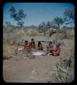 Group of men sitting together; one working with mortar and pestle