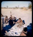 Food, Meat: Squatting man handing piece of cooked meat to another man, with other people watching them