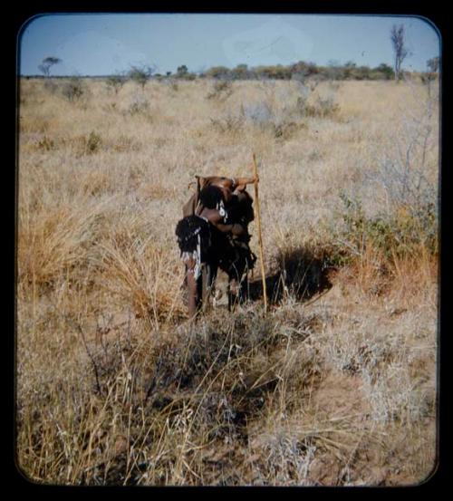 Food Gathering, Children: Woman carrying a child on her back, leaning over to pick something up; child is holding a stick