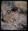 Food Gathering, General: Woman with a baby on her back, squatting and holding stem of root