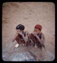 Greeting gesture: Two boys squatting with their hands to their faces; one with a bandaged foot and the other with ochred hair