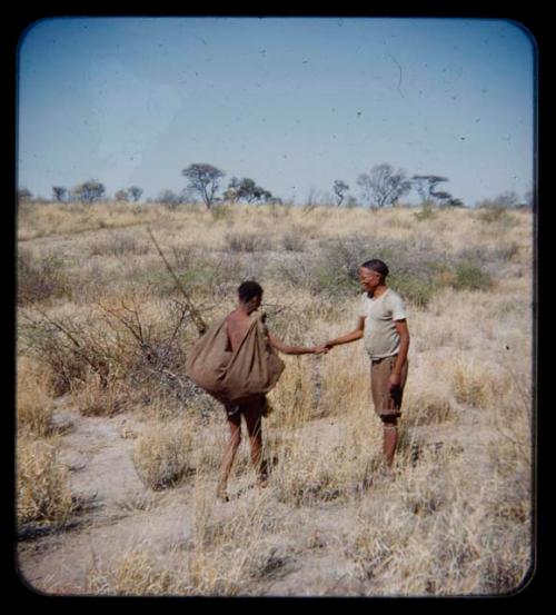 Groups: Man wearing T-shirt and trousers shaking hands with a man carrying kaross on his back