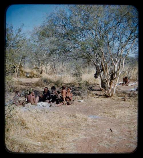 Groups: Group of people sitting around ashes of fire, with a man working in the background