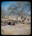 Groups: Group of people sitting around ashes of fire, with a man working in the background