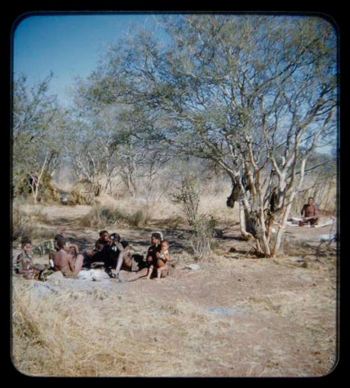 Groups: Group of people sitting around ashes of fire, with a man working in the background