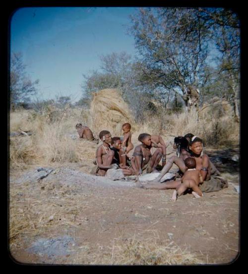 Groups: Group of people sitting around ashes of fire, with a man working in the background