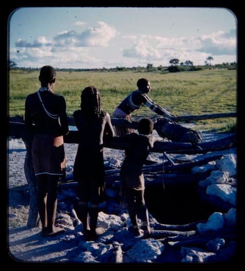 Makiena (wife of Muremi) standing and getting water at a waterhole, with Rungiero (her son's wife), Rungiero's sister and a child watching her