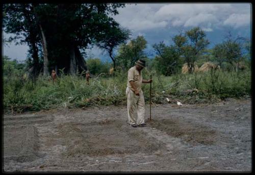 Wilhelm Camm standing with a stick in a cleared garden, with two men in the background