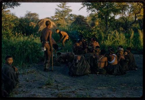 Group of people sitting and watching a curing dance, with huts and trees in the background