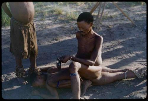 Medicine man sitting on /Gunda, who is lying on the ground with his face down