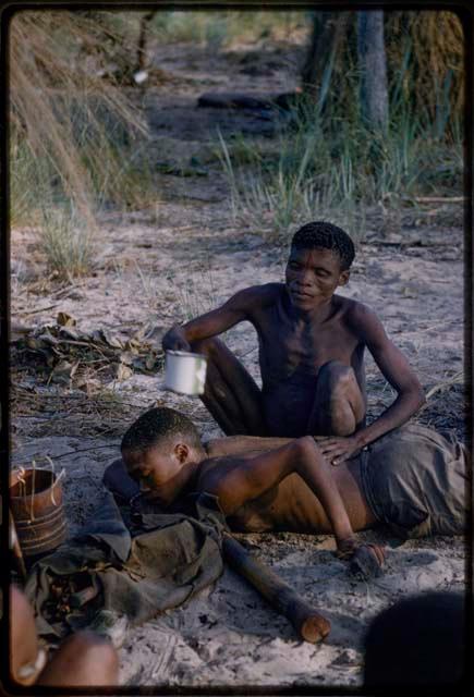 Medicine man "Short Kxao" holding an enamel cup over /Gunda lying on the ground with his face down