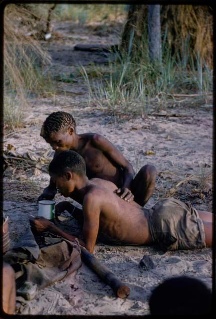 /Gunda looking at the content of an enamel cup handed over by medicine man "Short Kxao"