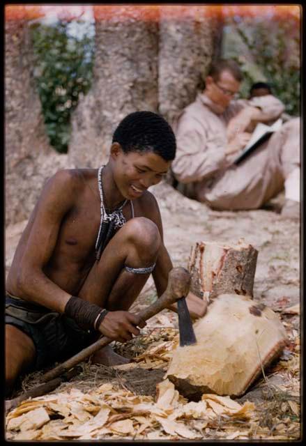 N!amshi chipping at log to make a //guashi, with Nicholas England in the background, leaning against a tree and reading