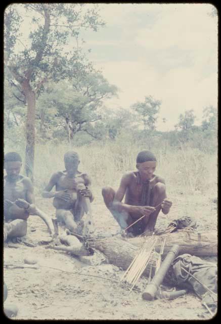 Kxao squatting and poisoning an arrow, with two men sitting nearby