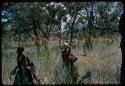 Two women walking in grass; one is carrying a bag and the other is holding an enamel bowl