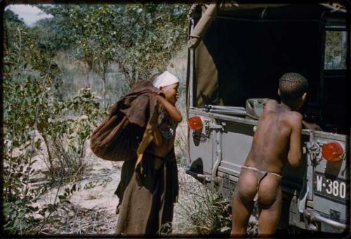 N!ai approaching Jeep, carrying a heavy bag of mangetti nuts over shoulder, with a boy standing nearby