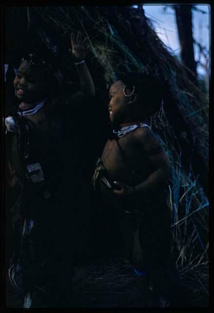 Two girls standing, with skerms in the background; one is the daughter of Hxua//a (one of Gaou's wives)