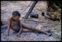 Boy sitting on the ground with nuts, with ≠Toma smoking in the background