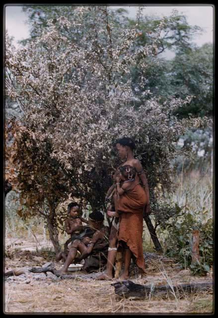 Two women with babies under a tree; one woman is wearing a red kaross