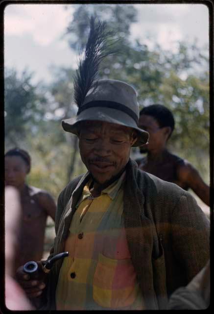 /Ti!kay standing and wearing Western clothes (jacket, plaid shirt, and felt hat with feather)