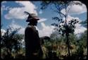 /Ti!kay in profile standing, wearing Western clothes (jacket, plaid shirt, and felt hat with feather), with two other men by tree in the background