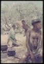 Tsamgao (≠Toma's son) sitting and playing a drum beat on a jerry can, with a man wearing Western clothes and a boy in the background