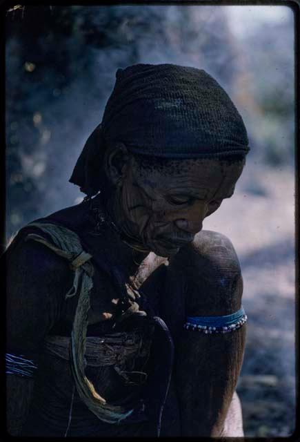 Kxao's wife in profile looking down, sitting on the ground