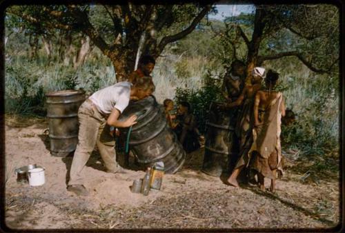 John Marshall siphoning from a camp water barrel, with other people (including ≠Toma and !U) watching him