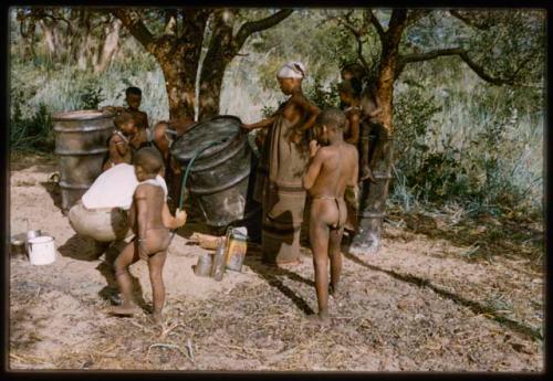 John Marshall siphoning from a camp water barrel while !U steadies it, with other people watching him