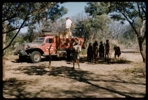 Robert Gardner photographing /Ti!kay's return from farms, on a red expedition truck reading "Peabody Museum of Harvard University," with expedition members and a group of people watching them 
