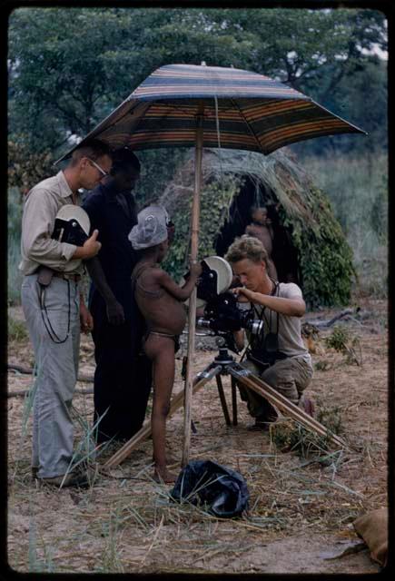 John Marshall checking a camera under an umbrella, with two men and a boy standing nearby and a skerm in the background